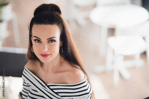 Beautiful woman with brunette hair, sexy smiling female in a cafe, blurred background. She has lovely smile. Stylish look, make up, light pink lipstick, casual outfit.