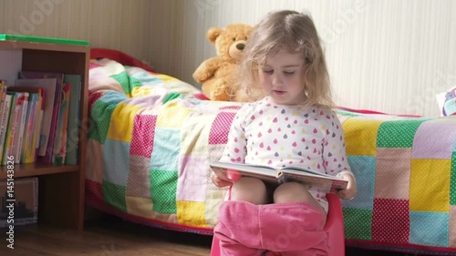 Little girl sitting on the potty beside the bed and reading a book.