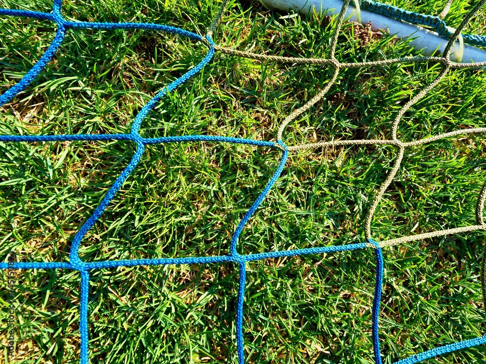 Detail of yellow blue crossed soccer nets, soccer football in goal net with grass on outdoor playground in the background. Detail of yellow blue crossed soccer nets, soccer football in goal net with grass on outdoor playground in the background.