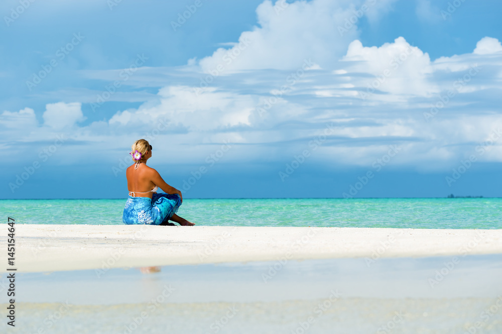 Blonde woman sitting on white sand beach, looking at open sea.Copy space