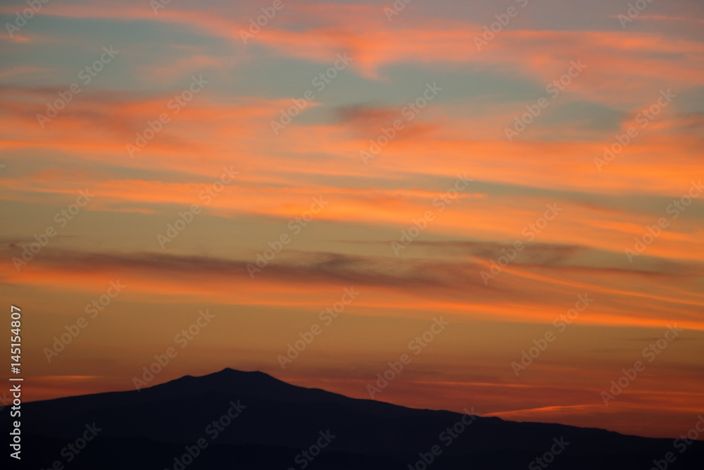 A top of mountain silhouette, under a big sky with beautiful, striped red clouds at sunset