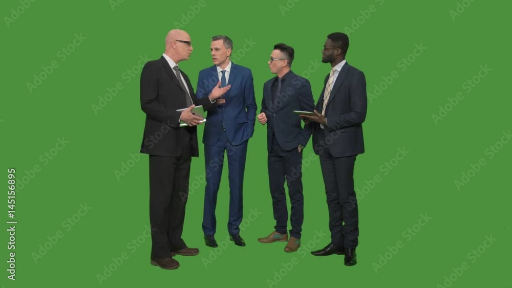 Four males in suits standing in circle and having discussion on green background using different gadgets tablet phones Four males in suits standing in circle and having discussion on green background using different gadgets tablet phones