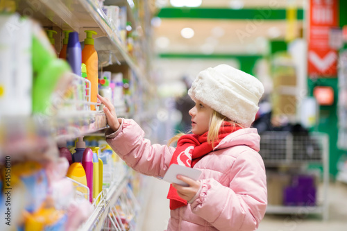Adorable girl in pink winter jacket and white fur hat selects cosmetics on shelf in supermarket