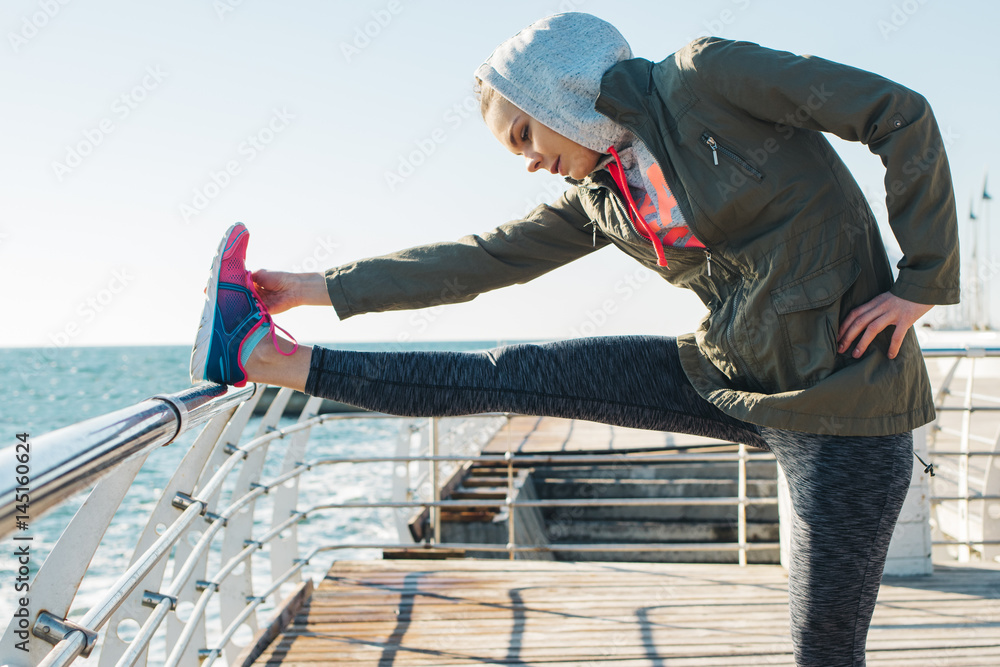 Young woman in jacket and hood stretches legs on the beach in the morning Young woman in jacket and hood stretches legs on the beach in the morning