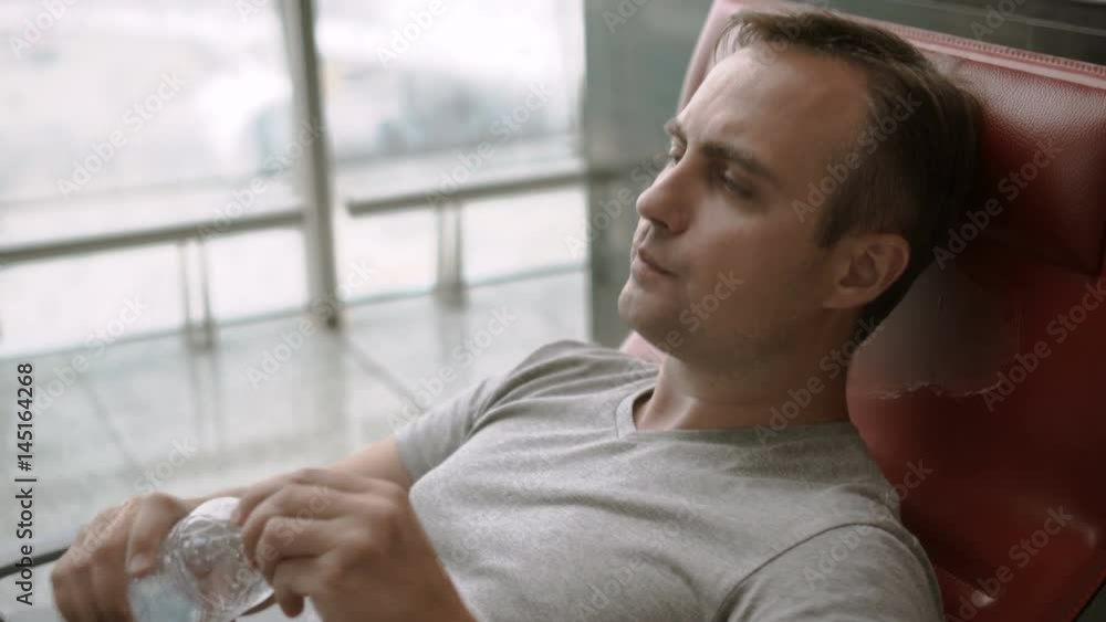 Handsome tired young man in airport terminal. Long distance connected flights jet-lag. Drinking water from plastic bottle Handsome tired young man in airport terminal. Long distance connected flights jet-lag. Drinking water from plastic bottle
