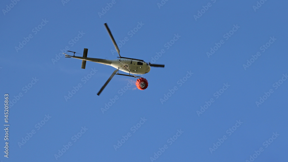 Firefighting helicopter with water bucket and trail of drops seen from below against blue sky Firefighting helicopter with water bucket and trail of drops seen from below against blue sky
