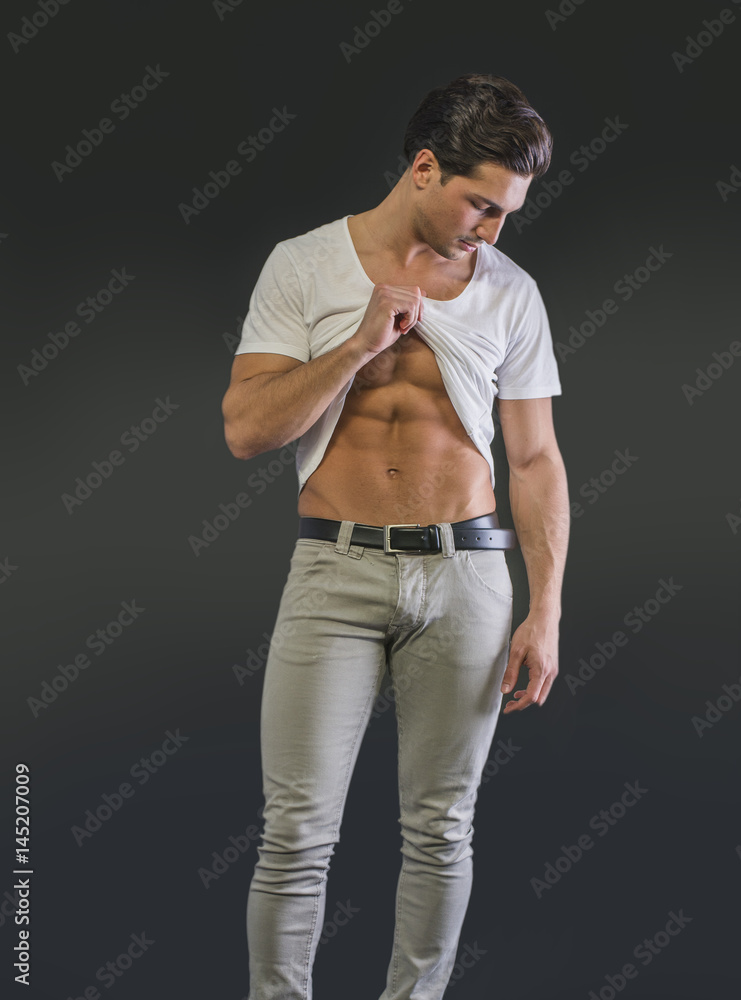Handsome young man pulling up t-shirt, showing abs, on dark background in studio Handsome young man pulling up t-shirt, showing abs, on dark background in studio