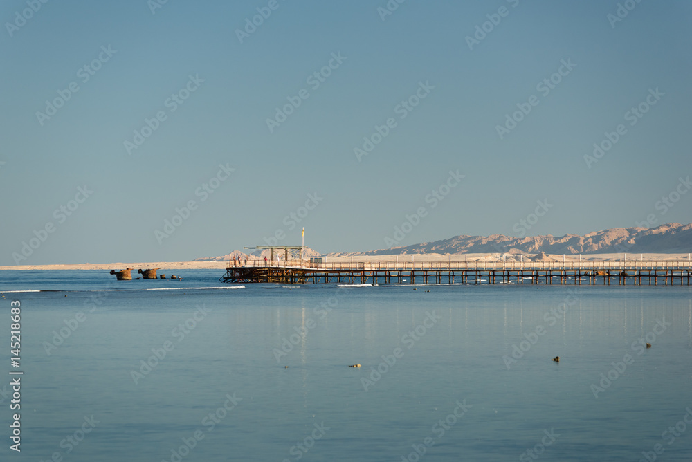 View at the distant pier of a tourist resort. Summer vacation on Red sea. View at a clear sea with turquoise water in windless conditions. Summer vacation at a sea coastline in an exotic country.