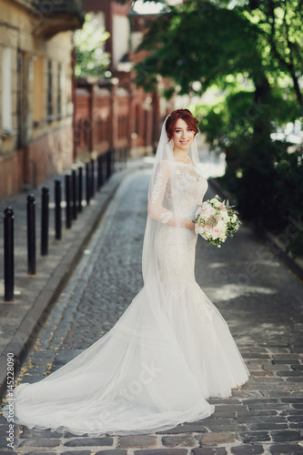 Bride with red hair holds white wedding bouquet standing on the street