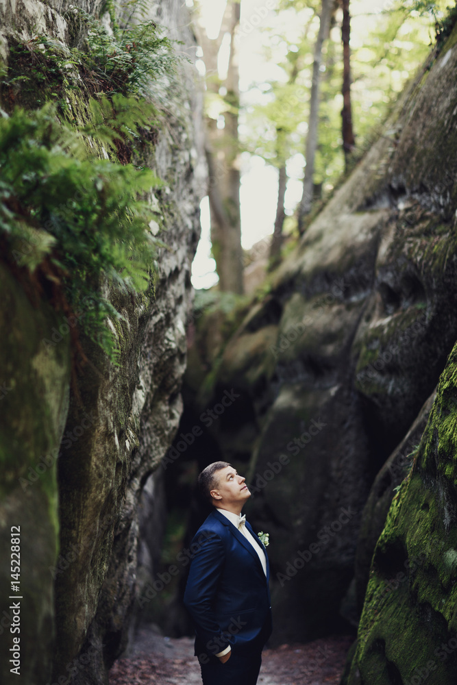 Handsome groom in blue suit poses between the rocks