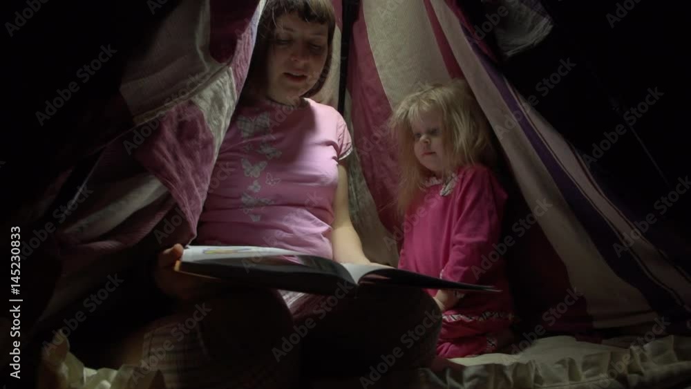 Mother and daughter in pink pajamas sit in a striped tent on the floor in a dark room at night. Tired woman reads the book to a joyful little girl in lodge before with flashlights going to bed. Mother and daughter in pink pajamas sit in a striped tent on the floor in a dark room at night. Tired woman reads the book to a joyful little girl in lodge before with flashlights going to bed.