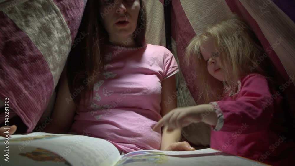Mother and daughter in pink pajamas sit in a striped tent on the floor in a dark room at night. Tired woman reads the book to a joyful little girl in lodge before with flashlights going to bed. Mother and daughter in pink pajamas sit in a striped tent on the floor in a dark room at night. Tired woman reads the book to a joyful little girl in lodge before with flashlights going to bed.
