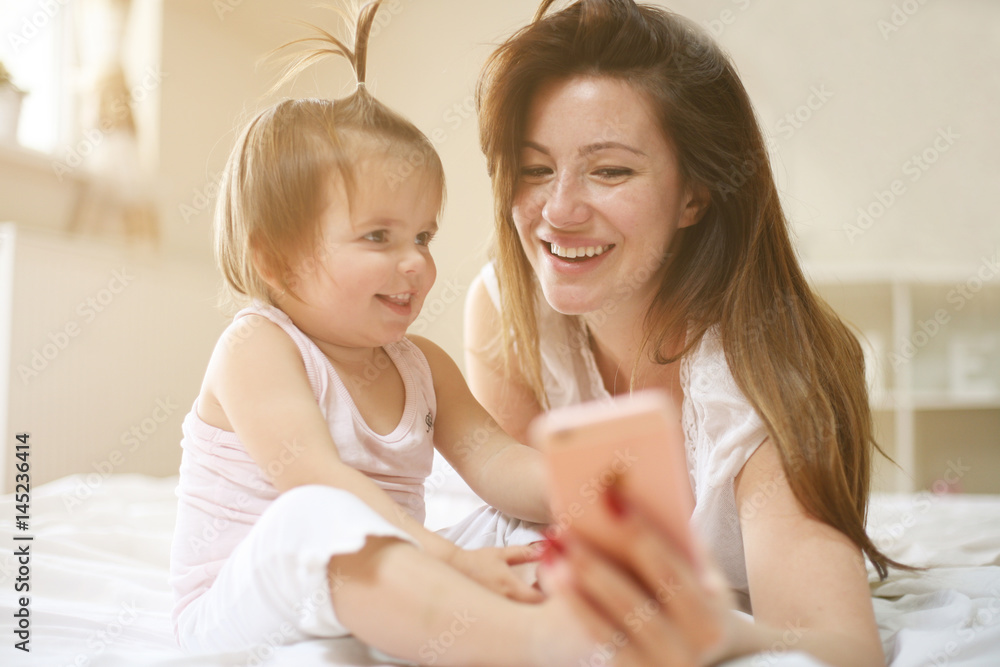 Mother with her little baby having fun in the bed. Using smart phone. Mother with her little baby having fun in the bed. Using smart phone.