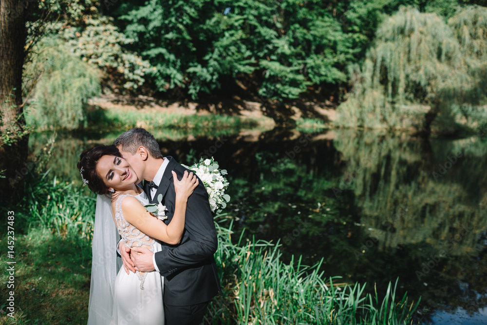 Groom kisses bride's neck standing with her before lake in park Groom kisses bride's neck standing with her before lake in park