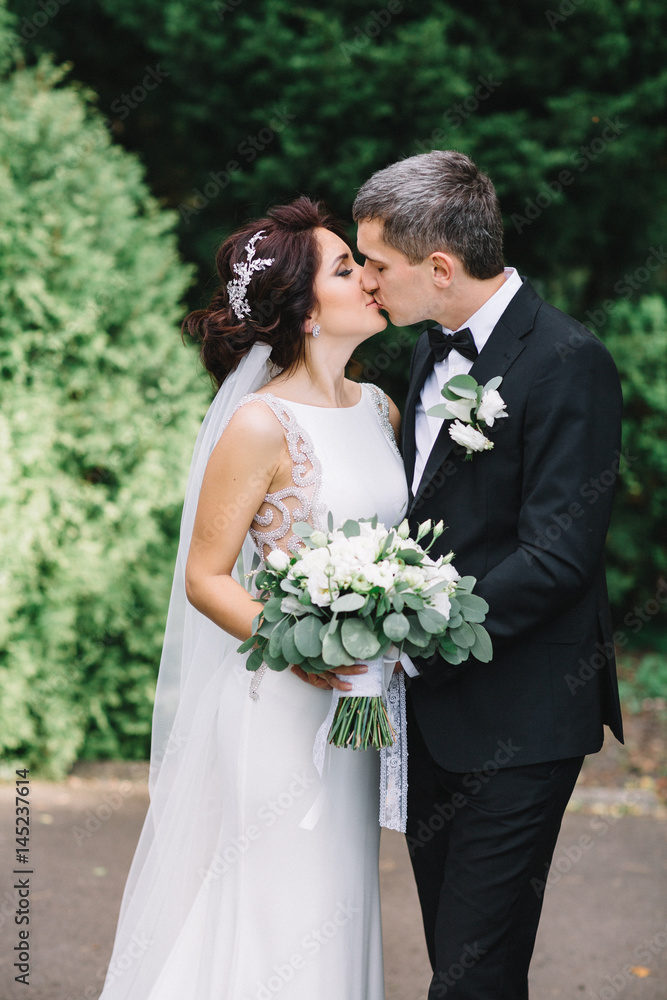 Tender kiss of bride and groom standing in green park Tender kiss of bride and groom standing in green park