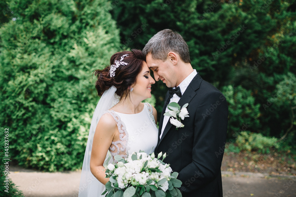 Bride leans to groom's forehead tender while he hugs her in the park Bride leans to groom's forehead tender while he hugs her in the park