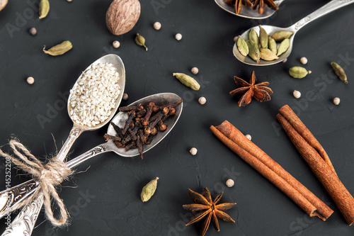 Various spices in metal vintage spoons and cinnamon sticks on dark rustic background. Cardamom, white pepper, nutmeg, anise, clove, sesame seeds. Aromatic food cooking and baking ingredients.
