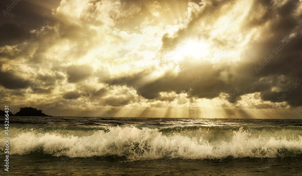 Dramatic cloudscape over the sea with rays of sunlight and a foaming wave in the foreground, toned warm colors
