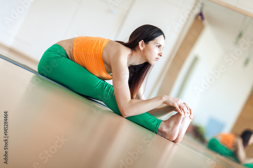 Sporty beautiful young woman in sportswear working out indoors, doing exercise for spine, shoulders, hamstrings, sitting in paschimothanasana pose, seated forward bend posture