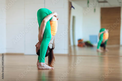 Young attractive woman practicing yoga, standing in head to knees exercise, uttanasana pose, working out wearing sportswear, indoor full length photo