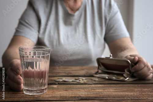 Senior woman sitting at table with glass of water, purse and coins, closeup. Poverty concept