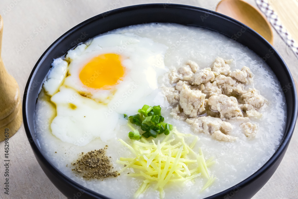Rice porridge is topping with ginger, spring onion, pepper, chicken, and onsen egg, Breakfast set on wooden table, spoon, pepper grinder. breakfast in Asia call Rice porridge is topping with ginger, spring onion, pepper, chicken, and onsen egg, Breakfast set on wooden table, spoon, pepper grinder. breakfast in Asia call