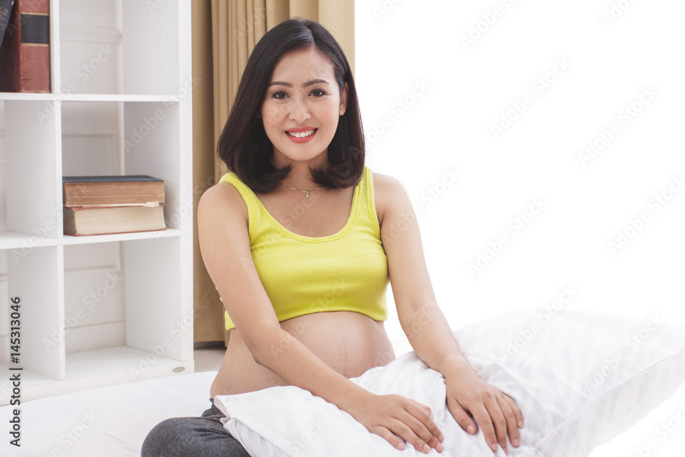 asian pregnant woman smiling while sitting on bed asian pregnant woman smiling while sitting on bed