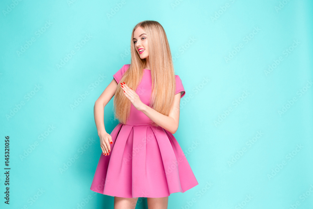 Fototapeta premium Smiling beautiful young woman in pink mini dress posing, presenting something and looking away. Three quarter length studio shot on yellow background.
