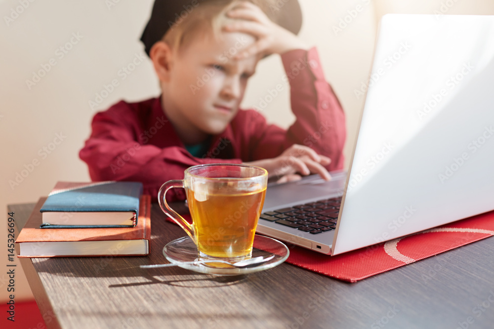 An emotional llitle boy wearing trendy clothes and cap sittiing at the table working with his laptop being tired of homework. A cup of tea in the foreground. A schoolboy preparing for the lessons. An emotional llitle boy wearing trendy clothes and cap sittiing at the table working with his laptop being tired of homework. A cup of tea in the foreground. A schoolboy preparing for the lessons.