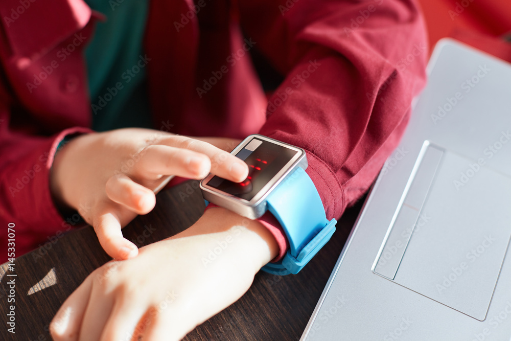 Obraz premium A close-up of child`s hands with smart watch. Touching electronic watch. Wearable gadget concept. Showing time. Using smartwatch while sitting near the laptop. Technology and people concept.