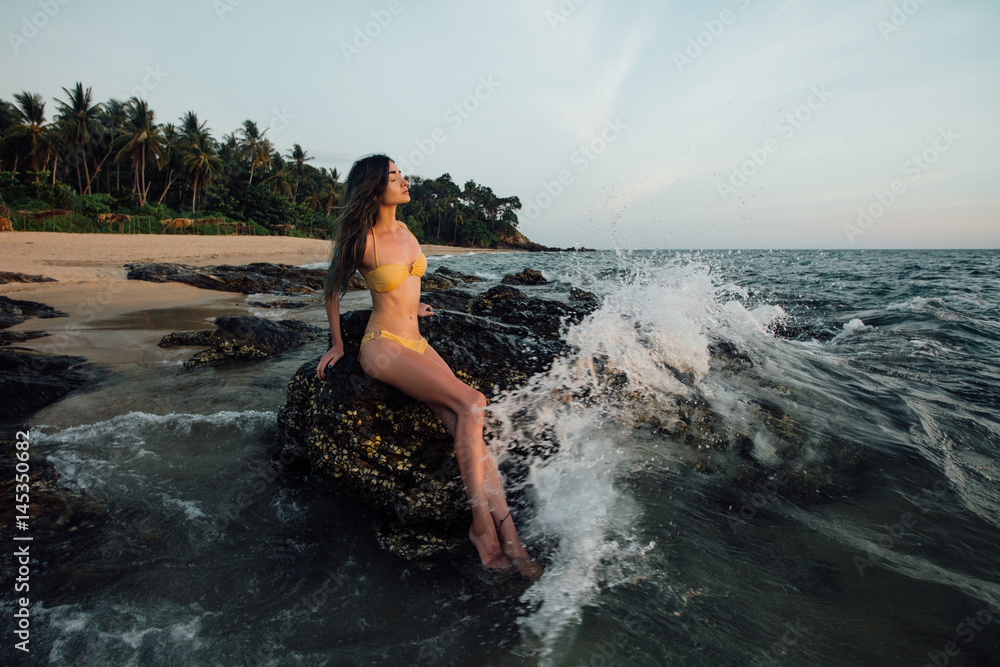 Sensual young brunette beauty wearing yellow swimsuit with beautiful hair sitting on stone at the beach