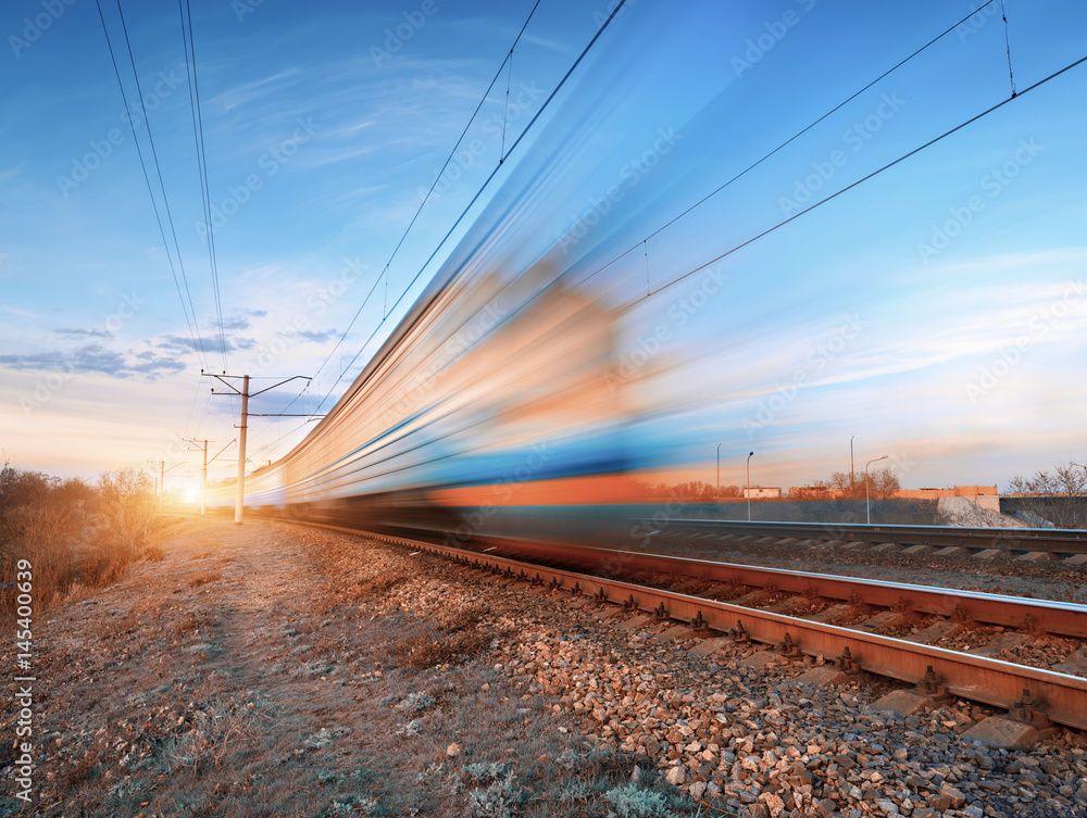 Obraz premium High speed train in motion on railroad track at sunset. Blurred commuter train. Railway station against colorful blue sky. Railroad travel, railway tourism. Rural industrial landscape in twilight