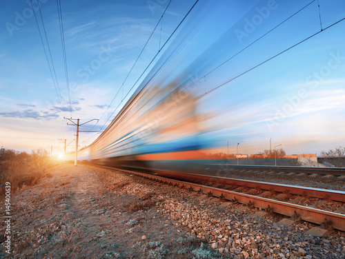 High speed train in motion on railroad track at sunset. Blurred commuter train. Railway station against colorful blue sky. Railroad travel, railway tourism. Rural industrial landscape in twilight