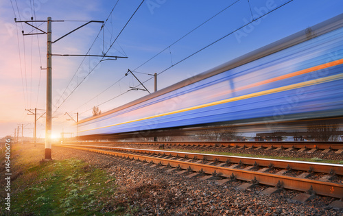 High speed passenger train in motion on railroad at sunset. Blurred commuter train. Railway station against colorful blue sky. Railroad travel, railway tourism. Rural industrial landscape. Concept