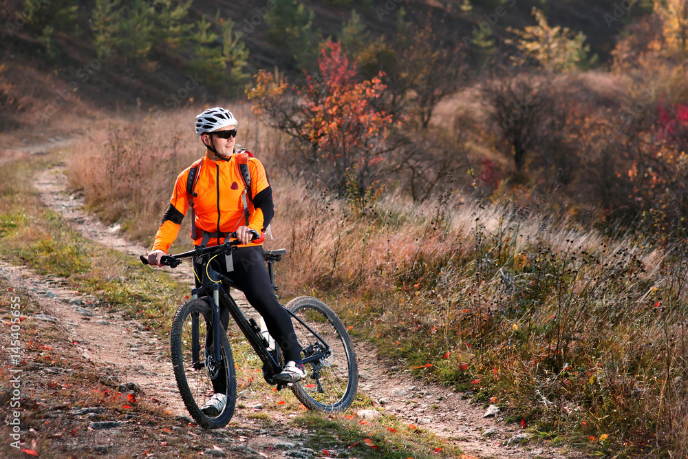 Naklejka premium Cyclist in the orange jacket riding a bike on countryside road at the sunrise.