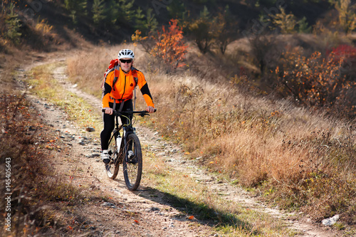 Cyclist in the orange jacket riding a bike on countryside road at the sunrise.