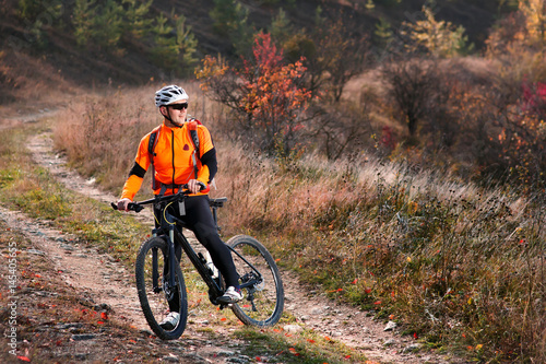 Cyclist in the orange jacket riding a bike on countryside road at the sunrise.