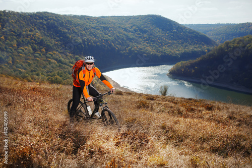 Cyclist in orange jacketr stands with his bike under river against beautiful landscape with mountain.