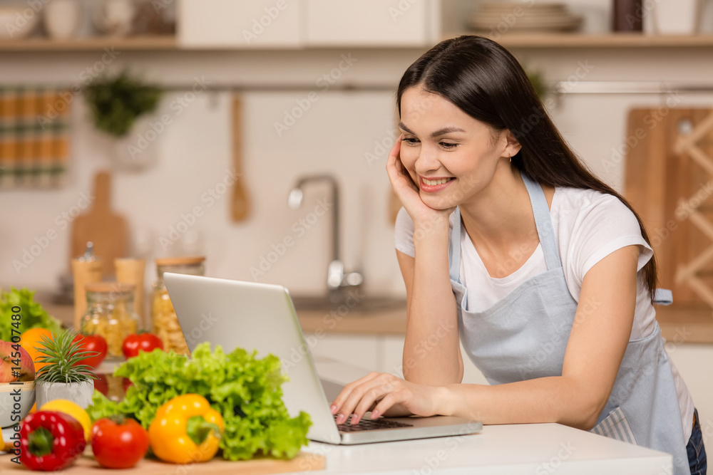 Young Woman standing in kitchen with tablet computer and looking recipes, smiling. Food blogger concept Young Woman standing in kitchen with tablet computer and looking recipes, smiling. Food blogger concept