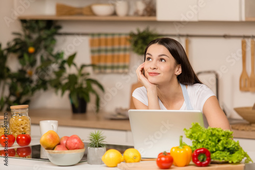Young woman standing near desk and laptop in the kitchen, smiling, dreaming. Food blogger concept