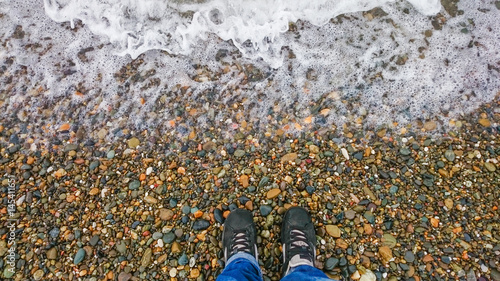 hipster in black shoes standing on a pebble beach in front of a receding wave of foam, curiosity concept