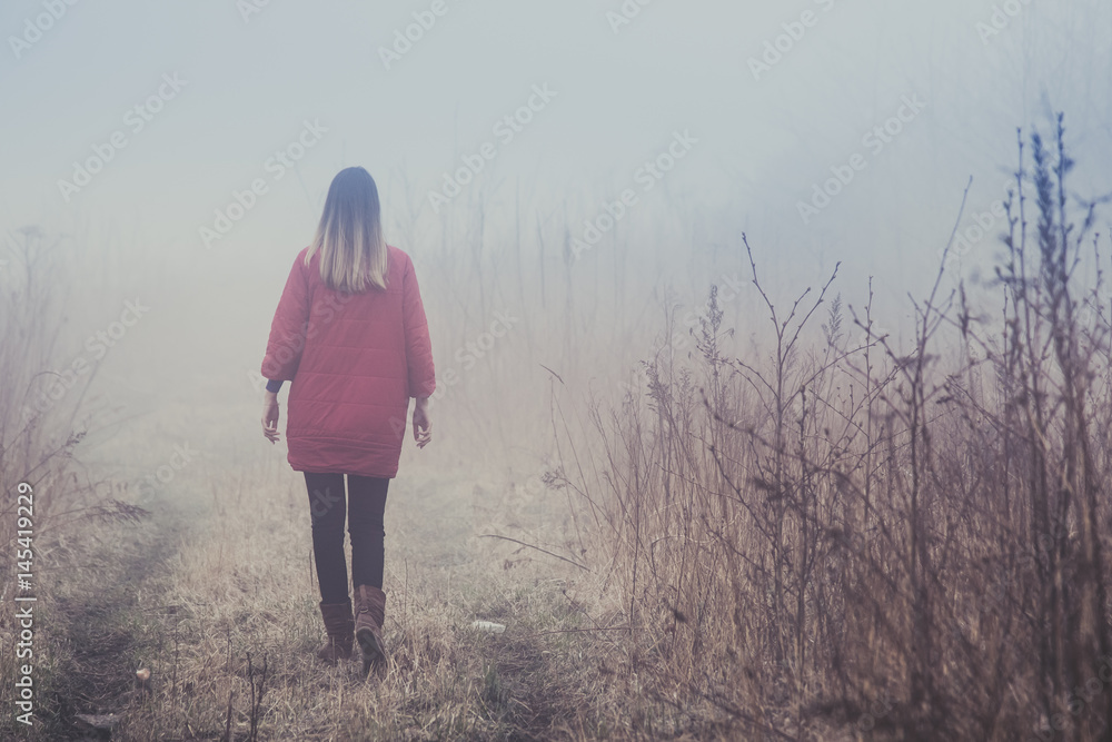 A girl in a red coat goes into the distance in a fog over a dry field