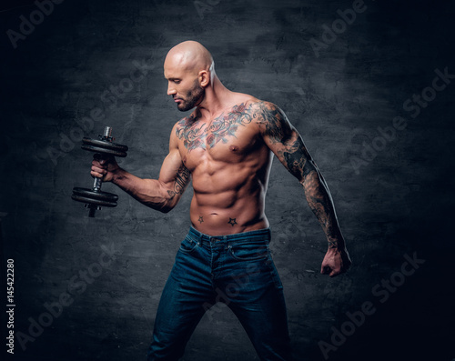 Shirtless shaved head, muscular male with tattoos on his chest and arms holds dumbbell. © Fxquadro