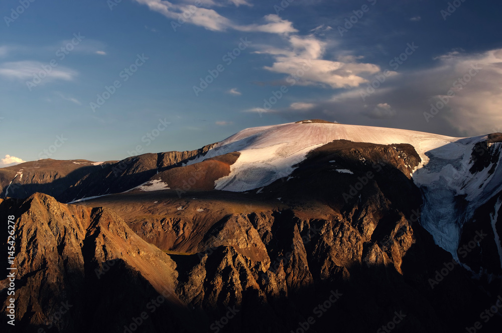 High mountain snow peak with glacier and dramatic rocks under sunset dawn blue sky and white clouds. Aktru, Altai Mountains, Siberia, Russia