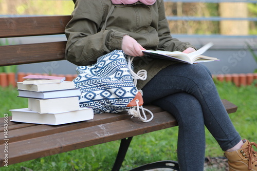 Młoda dziewczyna, uczennica, w zielonej kurtce, jeansowych spodniach, siedzi na ławce w parku, na kolanach trzyma rozłożona książkę, obok niej na ławce leży młodzieżowa torba i stos książek 