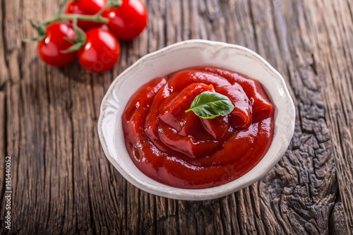 Ketchup or tomato sauce in white bowl and cherry tomatoes on wooden table.
