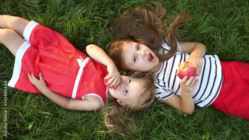 Two little sister girls eating apples lying on green grass in a park. Have a bite in the open air. Family picnic. Happy childhood. Two little sister girls eating apples lying on green grass in a park. Have a bite in the open air. Family picnic. Happy childhood.