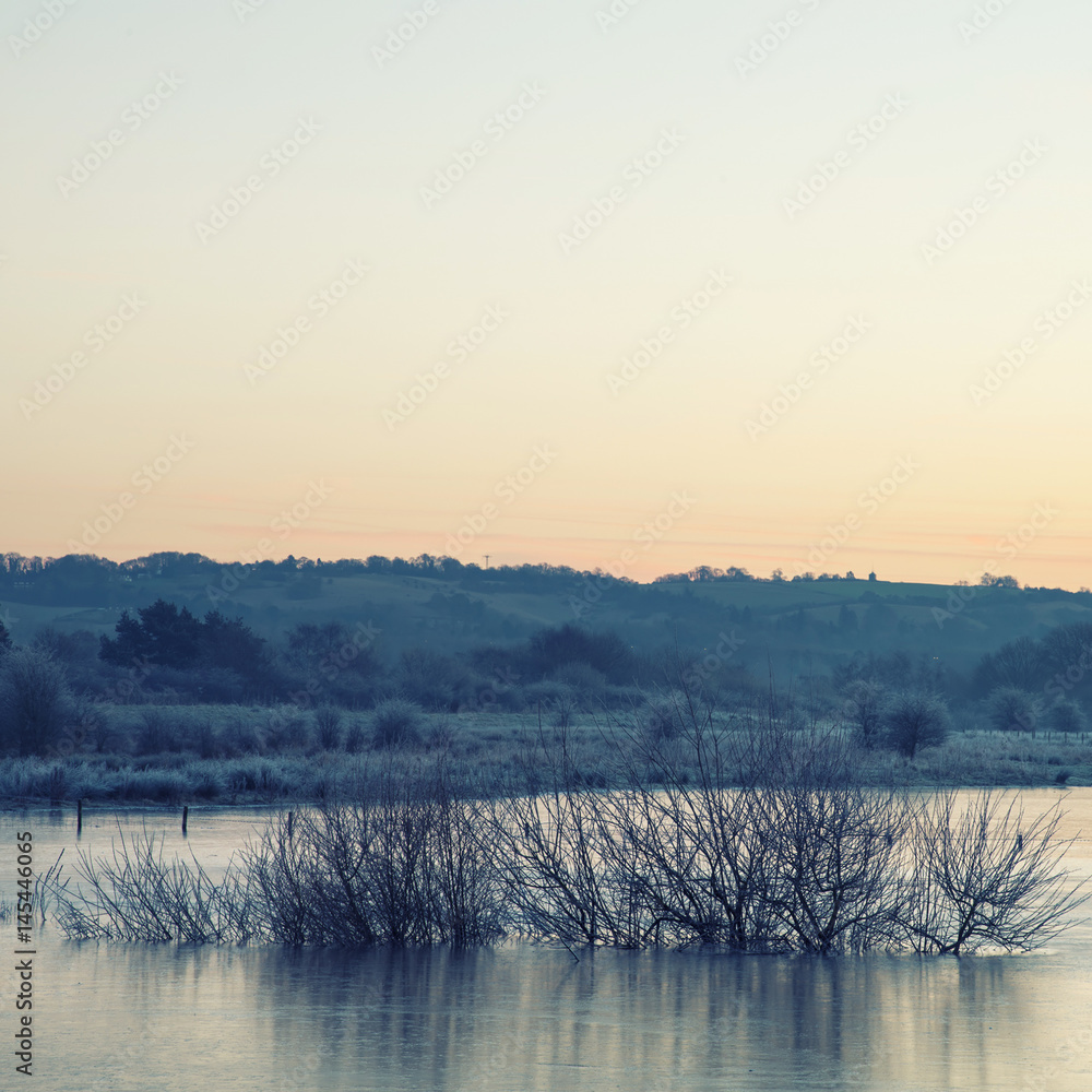 Beautiful vibrant English countryside lake image with frost and frozen lake in Winter at sunrise