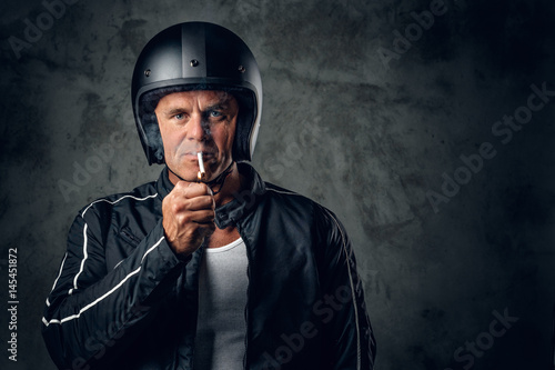 A man in motorcycle leather jacket and safety helmet smokes a cigarette.