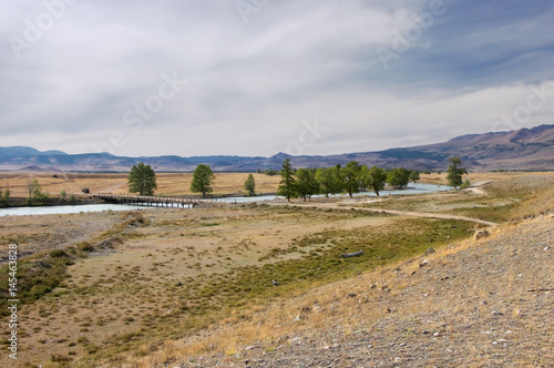 Log wooden bridge across the mountain river at steppe with dry yellow grass on a background of hills under cloudy sky Kurai Altai Mountains, Siberia, Russia